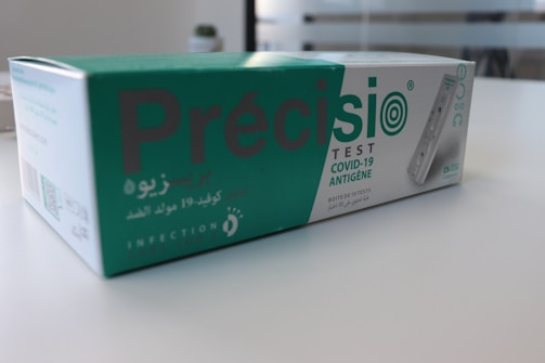 A box containing a COVID-19 antigen test kit, labeled 'Précisio Test', is placed on a white surface. The box is primarily green and white, with text in both English and Arabic. The image shows the product design and branding details.