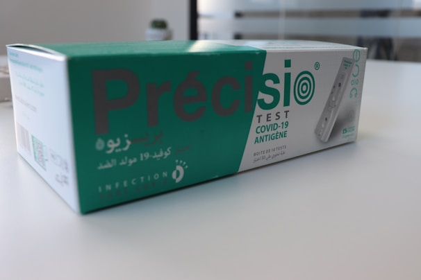 A box containing a COVID-19 antigen test kit, labeled 'Précisio Test', is placed on a white surface. The box is primarily green and white, with text in both English and Arabic. The image shows the product design and branding details.