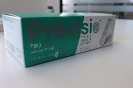 A box containing a COVID-19 antigen test kit, labeled 'Pr&eacute;cisio Test', is placed on a white surface. The box is primarily green and white, with text in both English and Arabic. The image shows the product design and branding details.