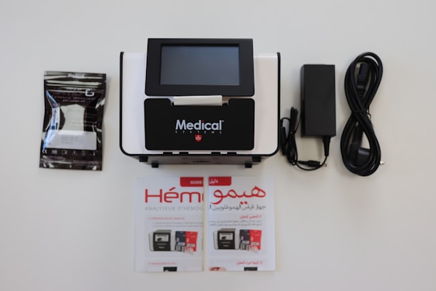 A medical device with a digital screen is centrally positioned. It is surrounded by a booklet, a package of medical supplies, and a power cable with an adapter. The booklet and package feature text.