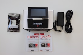 A medical device with a digital screen is centrally positioned. It is surrounded by a booklet, a package of medical supplies, and a power cable with an adapter. The booklet and package feature text.