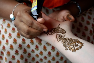 A person is applying intricate henna designs on another person's arm using a henna cone. The design features floral and geometric patterns. The background shows fabric with a repeating floral pattern.