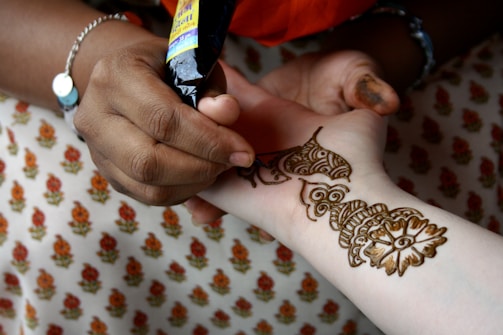 A person is applying intricate henna designs on another person's arm using a henna cone. The design features floral and geometric patterns. The background shows fabric with a repeating floral pattern.
