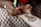A person is applying intricate henna designs on another person's arm using a henna cone. The design features floral and geometric patterns. The background shows fabric with a repeating floral pattern.