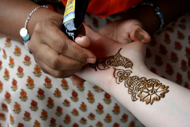 A person is applying intricate henna designs on another person's arm using a henna cone. The design features floral and geometric patterns. The background shows fabric with a repeating floral pattern.