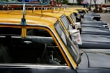 A line of classic black and yellow taxis is parked closely together on a street. Each taxi features a yellow roof and racks, with side mirrors and license plates visible. Advertisements in a different language are attached to the side mirrors. Trees and other vehicles can be seen in the background.