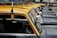 A line of classic black and yellow taxis is parked closely together on a street. Each taxi features a yellow roof and racks, with side mirrors and license plates visible. Advertisements in a different language are attached to the side mirrors. Trees and other vehicles can be seen in the background.