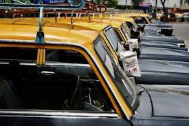 A line of classic black and yellow taxis is parked closely together on a street. Each taxi features a yellow roof and racks, with side mirrors and license plates visible. Advertisements in a different language are attached to the side mirrors. Trees and other vehicles can be seen in the background.