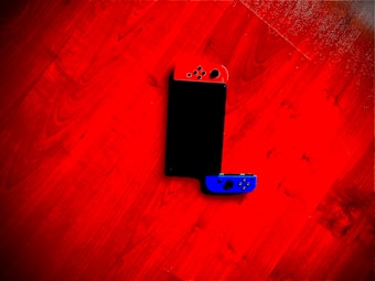 A gaming console with detachable red and blue controllers lies on a red wooden floor. The device is positioned diagonally with one controller partially removed.