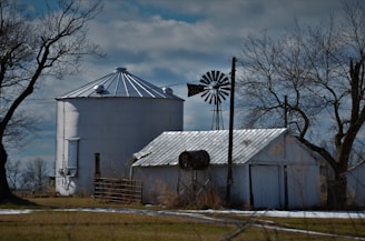 A rural scene features a large metallic grain silo flanked by a weathered barn and an old windmill. Leafless trees contrast against the cloudy sky, hinting at an early spring or late autumn setting. The landscape is predominantly flat with patches of bare ground and sparse grass.