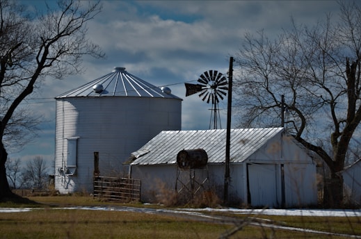 A rural scene features a large metallic grain silo flanked by a weathered barn and an old windmill. Leafless trees contrast against the cloudy sky, hinting at an early spring or late autumn setting. The landscape is predominantly flat with patches of bare ground and sparse grass.
