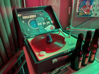 A retro-style turntable with a red vinyl record playing, accompanied by a vintage album cover featuring an illustrated space theme. Adjacent to the turntable are three dark glass bottles, possibly beer, with labels. In the background, there is a poster showing a person and the phrase '24 tattoos'. The setting has a nostalgic and vintage atmosphere, enhanced by dim lighting with green and red hues.