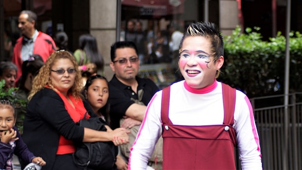 A performer with colorful face paint and expressive makeup is smiling with a crowd of people standing nearby. The performer is wearing a red costume with a white shirt underneath. The onlookers include men, women, and children, and they appear to be observing the performance with interest.