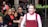A performer with colorful face paint and expressive makeup is smiling with a crowd of people standing nearby. The performer is wearing a red costume with a white shirt underneath. The onlookers include men, women, and children, and they appear to be observing the performance with interest.