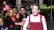 A performer with colorful face paint and expressive makeup is smiling with a crowd of people standing nearby. The performer is wearing a red costume with a white shirt underneath. The onlookers include men, women, and children, and they appear to be observing the performance with interest.