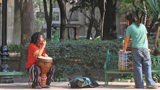 Two individuals with dreadlocks are in a park. One is seated on a green bench, playing a drum with colorful patterns, and wearing a red shirt and striped pants. The other person is standing near another green bench, interacting with a large drum placed on it. The scene takes place among trees and greenery, with a lamppost nearby.
