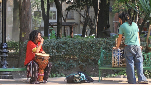 Two individuals with dreadlocks are in a park. One is seated on a green bench, playing a drum with colorful patterns, and wearing a red shirt and striped pants. The other person is standing near another green bench, interacting with a large drum placed on it. The scene takes place among trees and greenery, with a lamppost nearby.