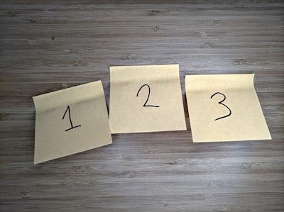 Three sticky notes are placed on a wooden surface, each with a number written in black marker. The notes are slightly curled at the top edges.