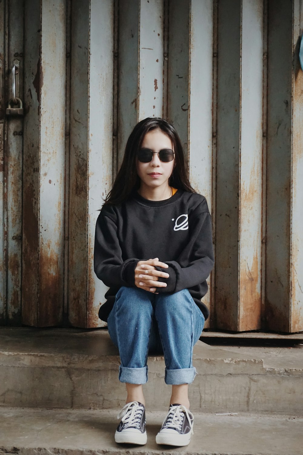 woman in black hoodie and blue denim jeans wearing black sunglasses sitting on brown wooden wall