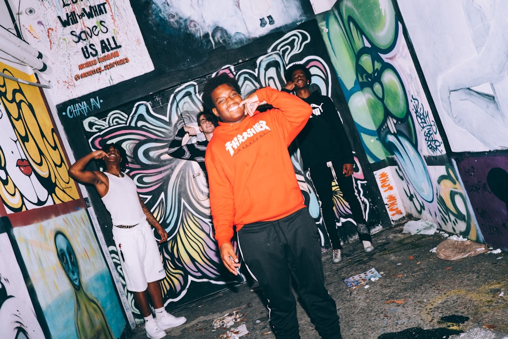 man in orange crew neck t-shirt and black pants standing beside graffiti wall during daytime