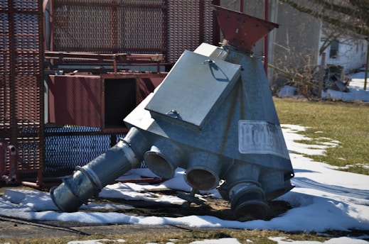 A large metallic industrial piece with multiple openings and a funnel top is situated on snow-covered ground in an outdoor setting. The metal structure appears weathered and is surrounded by machinery and a mesh-like fence.