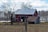 A red barn with a metal roof is situated in a rural landscape, surrounded by leafless trees and a grassy field. An old wooden shed with a rusted roof is adjacent to the barn. In the background, a line of bare trees stands against a partly cloudy sky.