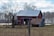 A red barn with a metal roof is situated in a rural landscape, surrounded by leafless trees and a grassy field. An old wooden shed with a rusted roof is adjacent to the barn. In the background, a line of bare trees stands against a partly cloudy sky.
