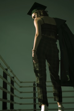 A person wearing a graduation cap and plaid trousers stands on a staircase, holding a dark graduation gown. The scene is captured from a low angle, emphasizing the vertical lines of the railing and the soft, dim lighting.