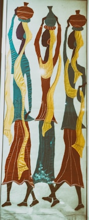 The image features a textile artwork depicting three stylized figures carrying vessels on their heads. The figures are elongated and abstract, with flowing garments in vibrant colors. The use of contrasting shades and seamless lines creates a sense of movement and harmony.
