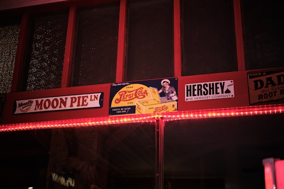 A nostalgic display featuring retro-style advertisements for Moon Pie, Pepsi-Cola, and Hershey. The signs are mounted on a wooden frame with a warm incandescent light strip running beneath. The vintage aesthetic is conveyed through classic logos and an old-fashioned design style.
