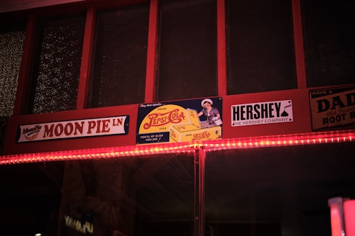 A nostalgic display featuring retro-style advertisements for Moon Pie, Pepsi-Cola, and Hershey. The signs are mounted on a wooden frame with a warm incandescent light strip running beneath. The vintage aesthetic is conveyed through classic logos and an old-fashioned design style.