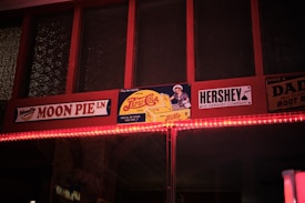 A nostalgic display featuring retro-style advertisements for Moon Pie, Pepsi-Cola, and Hershey. The signs are mounted on a wooden frame with a warm incandescent light strip running beneath. The vintage aesthetic is conveyed through classic logos and an old-fashioned design style.
