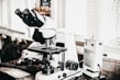 A laboratory setting features a high-powered microscope with several lenses and adjustment knobs placed on a countertop. Nearby, there is a white device labeled 'Quick Fix' alongside other laboratory equipment. The setting appears orderly, illuminated by natural light from a window with blinds in the background.