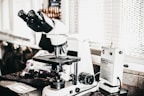 A laboratory setting features a high-powered microscope with several lenses and adjustment knobs placed on a countertop. Nearby, there is a white device labeled 'Quick Fix' alongside other laboratory equipment. The setting appears orderly, illuminated by natural light from a window with blinds in the background.