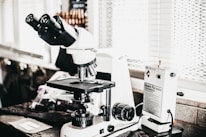 A laboratory setting features a high-powered microscope with several lenses and adjustment knobs placed on a countertop. Nearby, there is a white device labeled 'Quick Fix' alongside other laboratory equipment. The setting appears orderly, illuminated by natural light from a window with blinds in the background.