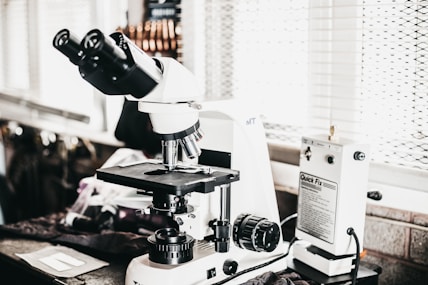 A laboratory setting features a high-powered microscope with several lenses and adjustment knobs placed on a countertop. Nearby, there is a white device labeled 'Quick Fix' alongside other laboratory equipment. The setting appears orderly, illuminated by natural light from a window with blinds in the background.