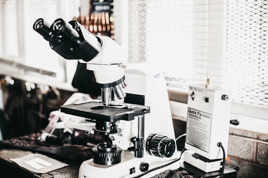 A laboratory setting features a high-powered microscope with several lenses and adjustment knobs placed on a countertop. Nearby, there is a white device labeled 'Quick Fix' alongside other laboratory equipment. The setting appears orderly, illuminated by natural light from a window with blinds in the background.
