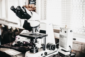 A laboratory setting features a high-powered microscope with several lenses and adjustment knobs placed on a countertop. Nearby, there is a white device labeled 'Quick Fix' alongside other laboratory equipment. The setting appears orderly, illuminated by natural light from a window with blinds in the background.