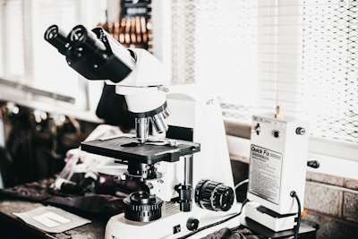 A laboratory setting features a high-powered microscope with several lenses and adjustment knobs placed on a countertop. Nearby, there is a white device labeled 'Quick Fix' alongside other laboratory equipment. The setting appears orderly, illuminated by natural light from a window with blinds in the background.