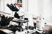 A laboratory setting features a high-powered microscope with several lenses and adjustment knobs placed on a countertop. Nearby, there is a white device labeled 'Quick Fix' alongside other laboratory equipment. The setting appears orderly, illuminated by natural light from a window with blinds in the background.