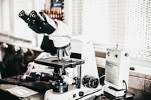 A laboratory setting features a high-powered microscope with several lenses and adjustment knobs placed on a countertop. Nearby, there is a white device labeled 'Quick Fix' alongside other laboratory equipment. The setting appears orderly, illuminated by natural light from a window with blinds in the background.