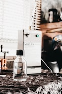 A small, clear bottle with a black cap is placed on a fabric-covered surface. In the background, a machine labeled 'Quick Fix' is visible, along with other workshop items and a window with blinds.
