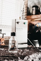 A small, clear bottle with a black cap is placed on a fabric-covered surface. In the background, a machine labeled 'Quick Fix' is visible, along with other workshop items and a window with blinds.