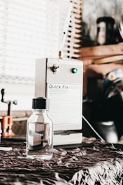 A small, clear bottle with a black cap is placed on a fabric-covered surface. In the background, a machine labeled 'Quick Fix' is visible, along with other workshop items and a window with blinds.