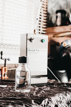A small, clear bottle with a black cap is placed on a fabric-covered surface. In the background, a machine labeled 'Quick Fix' is visible, along with other workshop items and a window with blinds.