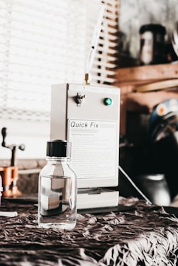 A small, clear bottle with a black cap is placed on a fabric-covered surface. In the background, a machine labeled 'Quick Fix' is visible, along with other workshop items and a window with blinds.