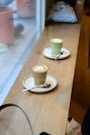 Two glasses of coffee sit on white saucers with spoons on a wooden countertop. One coffee is topped with latte art and the other appears to be a matcha latte. The setting is indoors, with natural light coming through a window.