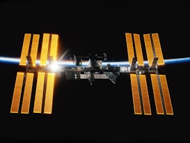 The image features the International Space Station (ISS) suspended against the backdrop of space. The solar panels are glowing a bright orange color, illuminated by the Sun's rays. A thin line of Earth's atmosphere is visible along the horizon, creating a stunning contrast between the stark darkness of space and the vibrant colors of the station and the Earth.