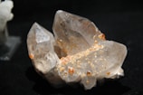 A cluster of clear quartz crystals with small, scattered orange or amber-colored inclusions on its surfaces, set against a dark background.