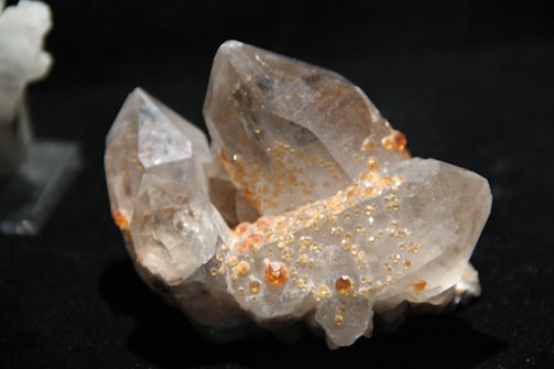 A cluster of clear quartz crystals with small, scattered orange or amber-colored inclusions on its surfaces, set against a dark background.